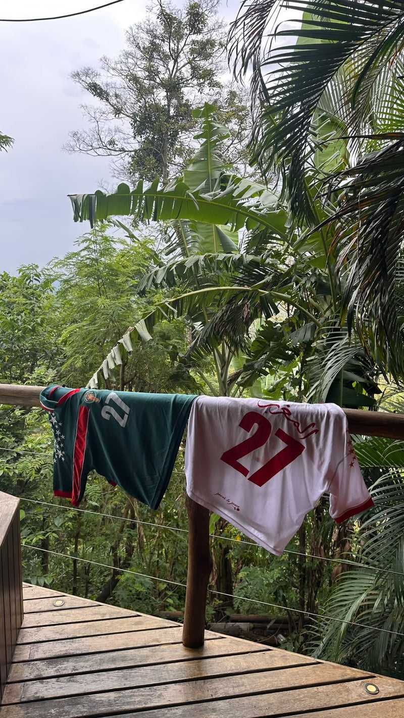 Les deux maillots REALAW DROP 01, vert et blanc et rouge, suspendus à une rambarde en bois devant la végétation