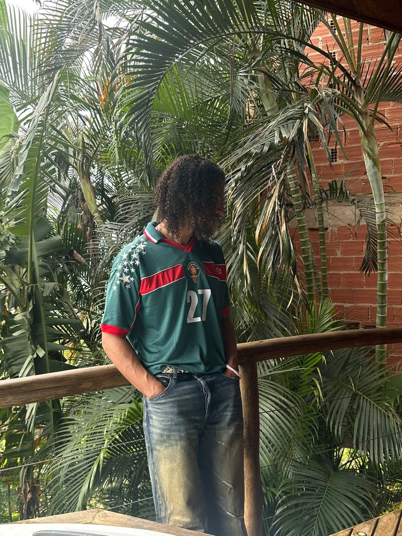 Personne en maillot REALAW DROP 01 vert, appuyée à une rambarde en bois au milieu des palmiers