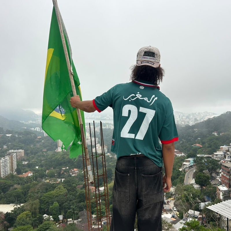 Personne vue de dos en maillot REALAW DROP 01 vert, numéro 27 et calligraphie arabe, un drapeau brésilien à la main au-dessus de la ville dans la brume